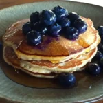 A stack of blueberry pancakes with fresh blueberries and honey on a rustic table.